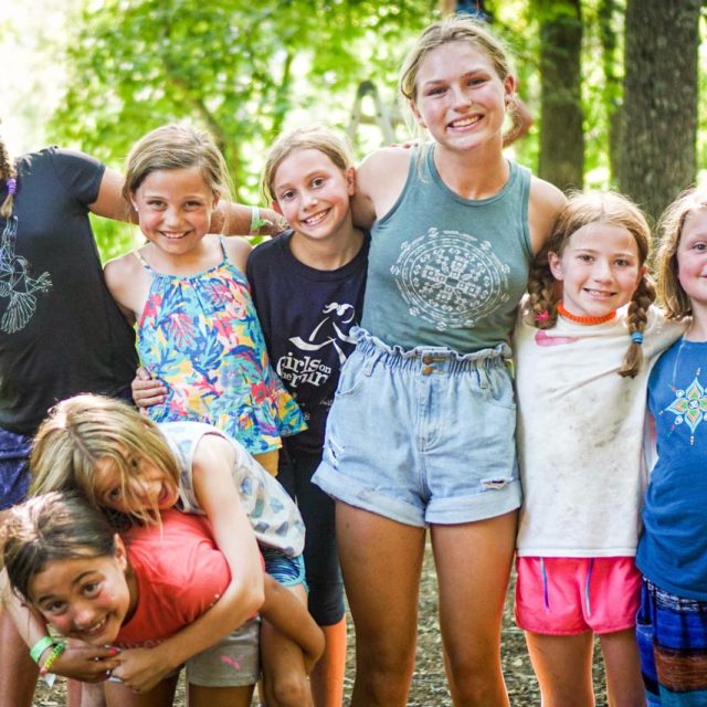 A group of eight children stands together outdoors in a wooded area at summer camp. They are smiling and posing closely, with some kids hugging each other and others having their arms around friends. The sunlight filters through the trees, illuminating their happy faces during summer camp.