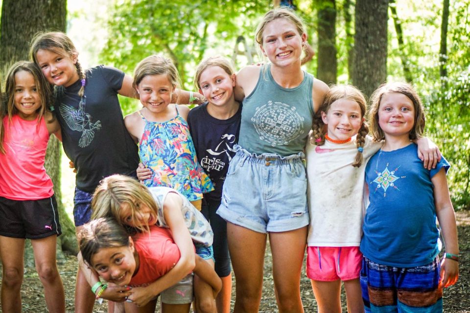 A group of eight children stands together outdoors in a wooded area at summer camp. They are smiling and posing closely, with some kids hugging each other and others having their arms around friends. The sunlight filters through the trees, illuminating their happy faces during summer camp.