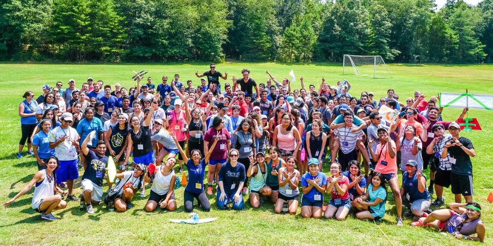 A large group of people, mostly young adults, are gathered on the sports field after a team building activity, smiling and posing for a photo. Many are wearing lanyards and casual summer clothing. There is a soccer goal in the background, and the surrounding area is filled with trees.