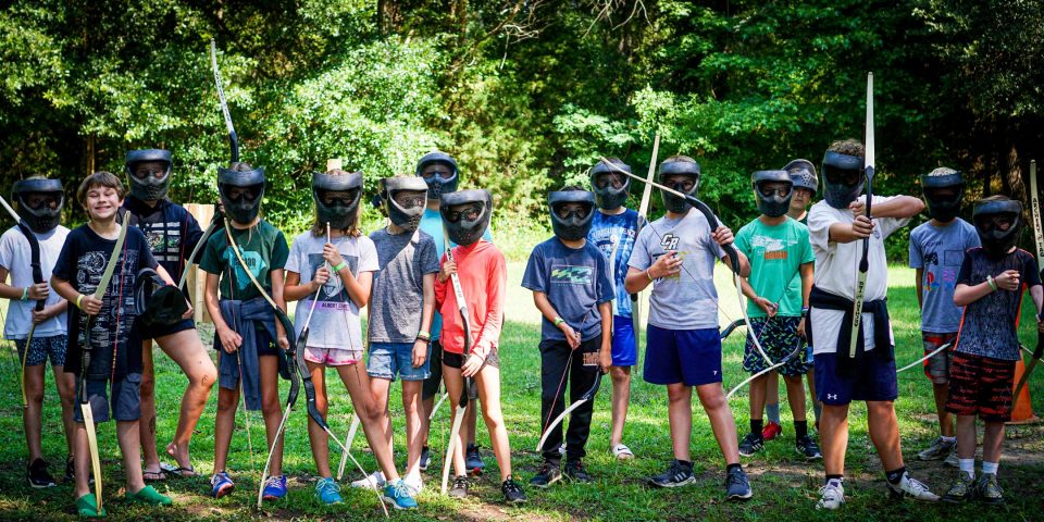 A group of children wearing protective masks and holding archery tag bows stand together outdoors on a grassy area with trees in the background.