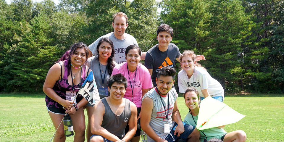 A group of nine young adults, wearing casual clothes and smiles, pose outdoors on a grassy field with trees in the background. Some hold kites and water bottles after completing a team building activity for their corporate outing. The sun is shining, creating a bright and cheerful atmosphere.