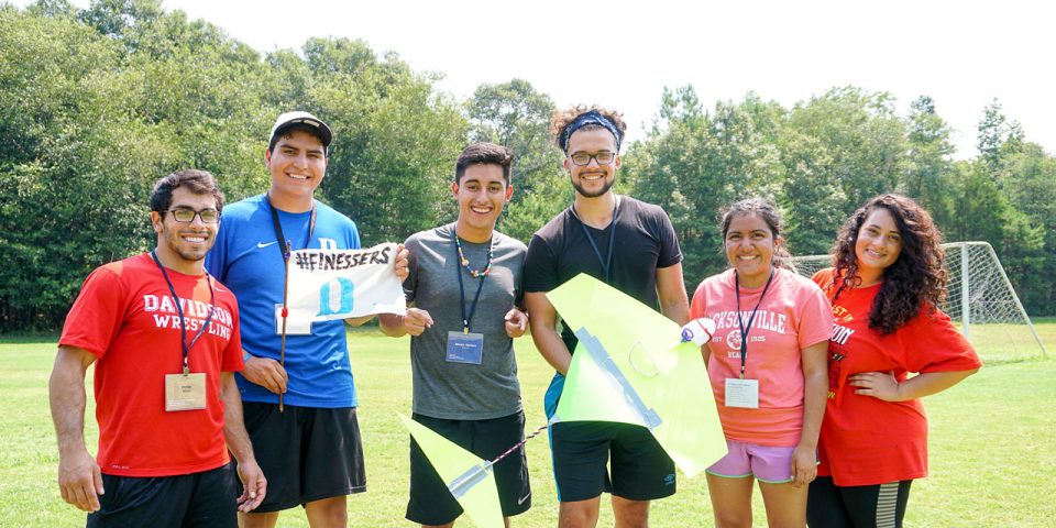 A group of six smiling people stand together outdoors on a sunny day at Camp Canaan. They are wearing casual clothing, with some dressed in red Davidson wrestling shirts. They hold up a colorful paper plane. Trees and a soccer goal are in the background.