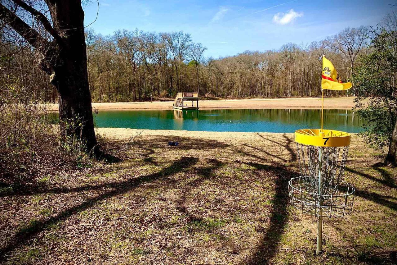 A disc golf basket with a yellow flag stands on the edge of a wooded area near a calm, clear lake at Camp Canaan. The sky is clear and blue, and a wooden double-decker dock is visible across the lake.