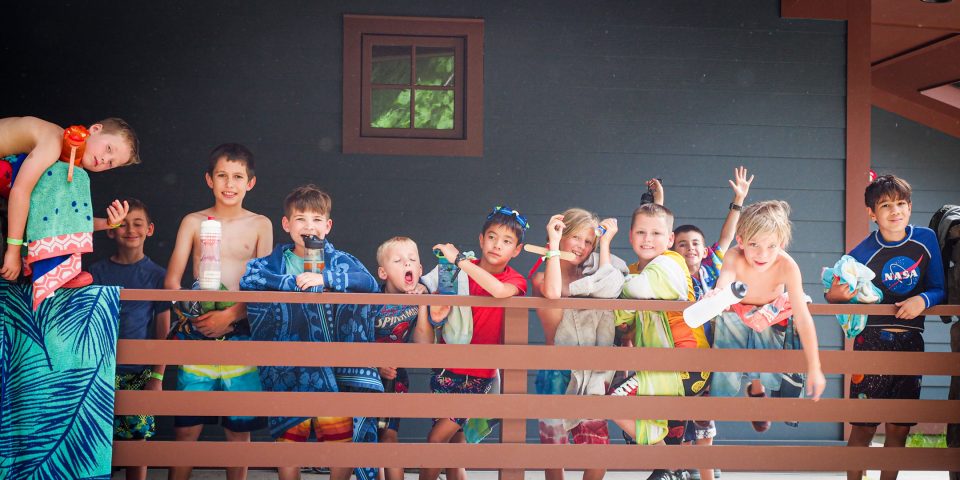 A group of children in swimwear stand outside of A&J Lodge during summer camp, smiling and making playful gestures toward the camera. Some hold towels, water bottles, or wear goggles. They appear excited and full of energy.