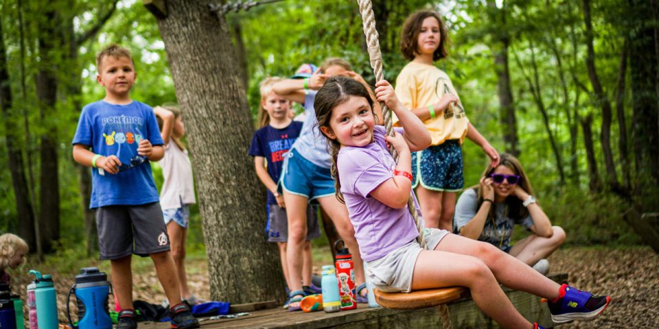 A group of children plays outdoors at Camp Canaan, a well-known summer camp in Rock Hill. One child, wearing a purple shirt and shorts, is swinging on a rope while others watch and wait their turn.