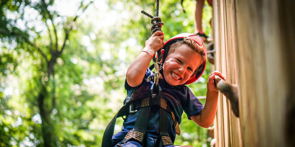 A young child wearing a red helmet and safety harness is smiling while climbing a rock climbing wall during summer camp.