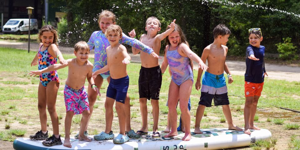 A group of children in swimsuits playfully pose on a large paddle board which is sitting on the ground by Lake Canaan.