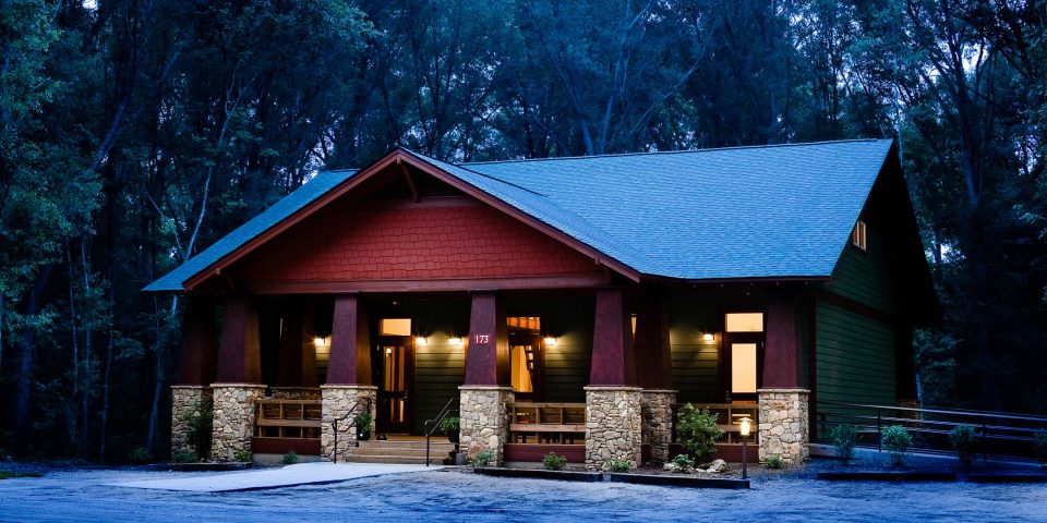 Exterior view of the multi-purpose building at Camp Canaan, with warm interior lights on stands, is surrounded by dense trees in a twilight setting. The building features a gabled roof, stone columns, and a front porch. The entrance is illuminated, creating a welcoming atmosphere perfect for corporate retreats amidst the forest backdrop.