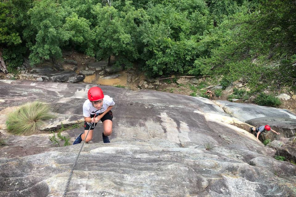 A camper wearing a red helmet and a white shirt climbs a rock face at Crowder's Mountain during summer camp.