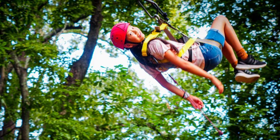 A girl wearing a red helmet and safety harness is ziplining through the woods at Camp Canaan. The individual, dressed casually in a light-colored shirt and shorts, enjoys the outdoor adventure amidst lush green trees, making the most of their summer camp experience.