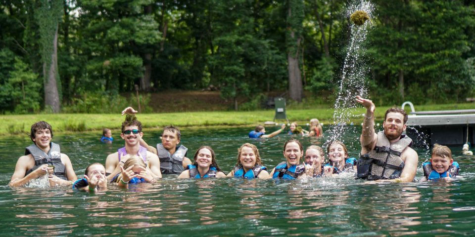 A group of people wearing life vests are enjoying a swim in Lake Canaan during summer camp. Some are splashing water and throwing objects, while others are smiling and posing for the photo. Trees and greenery surround the area.