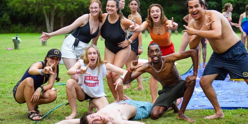 A group of happy counselors pose outdoors on the sports fields during summer camp. Some are wearing swimwear, and others are dressed casually. One person is lying on the ground, playfully posing. Trees and other relaxed individuals are visible in the background.