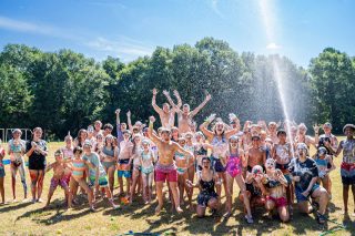 A large group of campers enjoy a spray of water from a hose held by a counselor on the sports field during summer camp. A soccer goal is in the background.