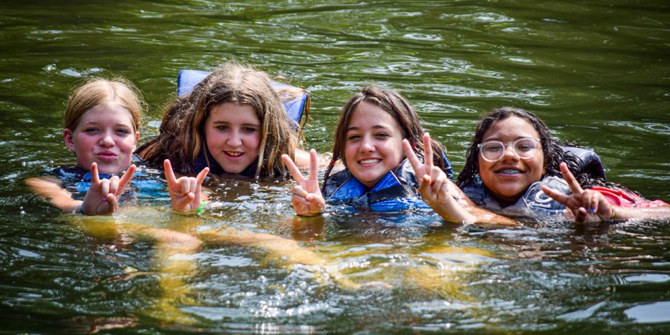 four girls give peace signs while floating in Lake Canaan with their life vests on