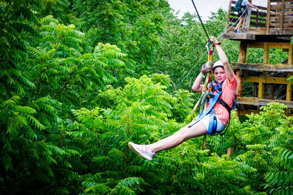 A girl wearing a red shirt, shorts, and a helmet is ziplining through the lush, green trees at Camp Canaan. They are harnessed and holding onto the zipline handle during their thrilling zipline canopy tour experience. A wooden platform is visible in the background.