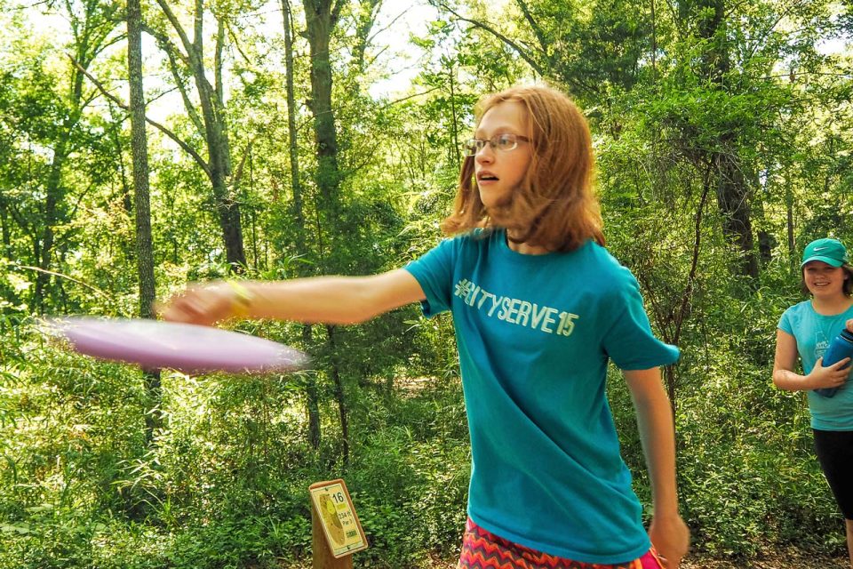 A young girl with glasses and reddish hair wearing a blue "CITY SERVE 15" T-shirt is throwing a pink disc while playing disc golf at Camp Canaan during summer camp.