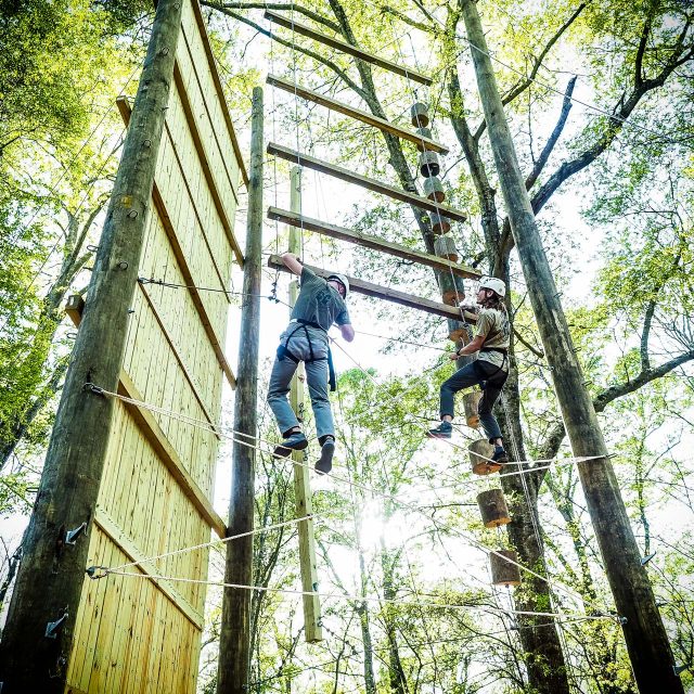 Two people wearing harnesses and helmets are climbing the Catawba Tower among trees at Camp Canaan. They navigate suspended wooden planks and ladders connected by ropes. Sunlight filters through the foliage, casting light on the climbers.