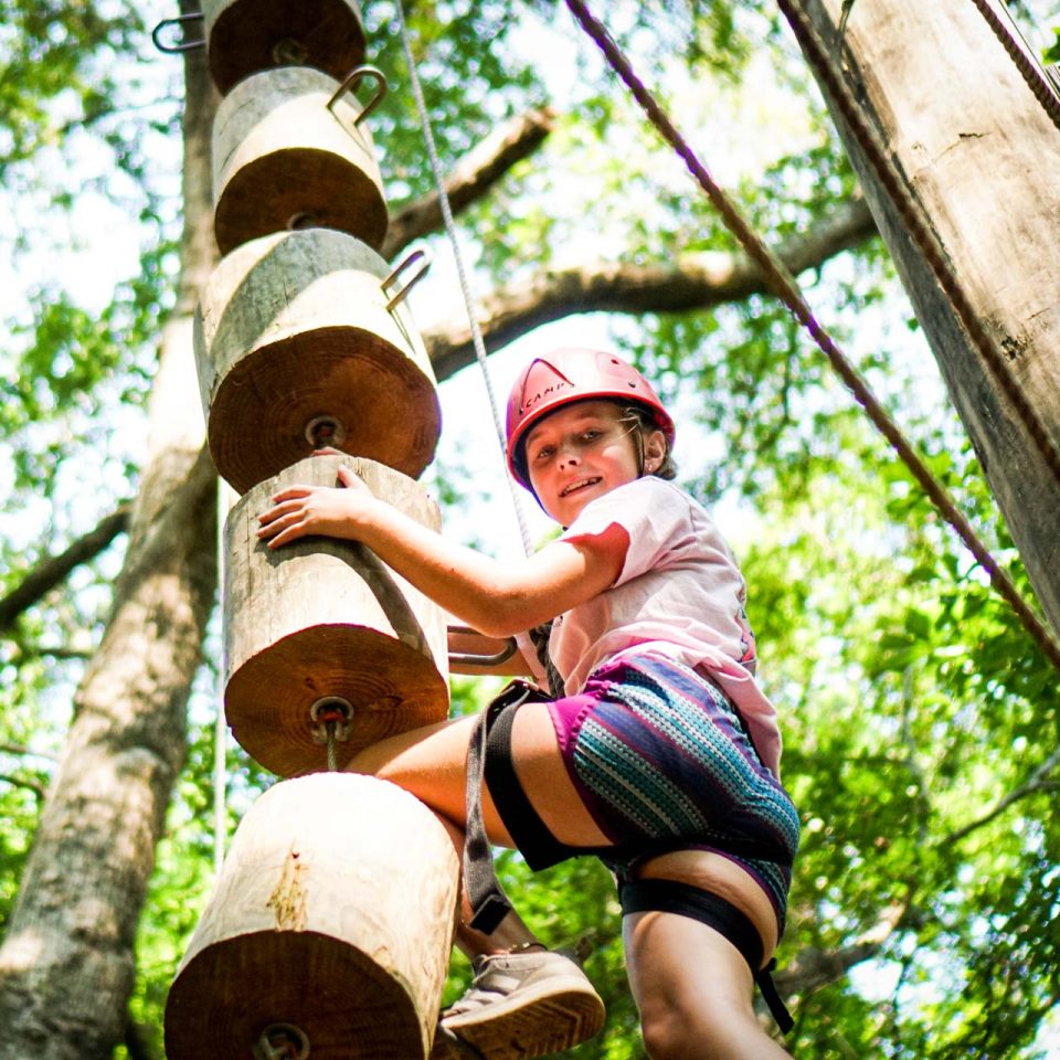 A young camper wearing a red helmet and safety harness is climbing a vertical wooden structure on the Catawba Tower at Camp Canaan during summer camp.