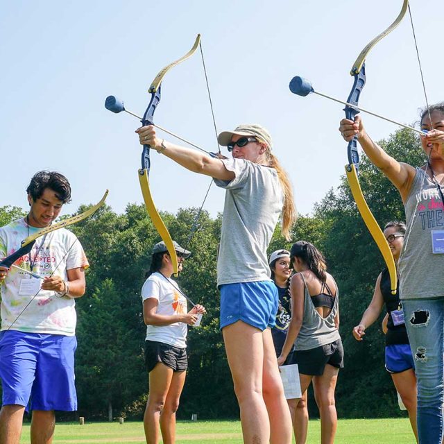 A group of people standing outdoors at a summer camp, holding archery tag bows and arrows. Two individuals are aiming their bows, while another looks down at his bow.