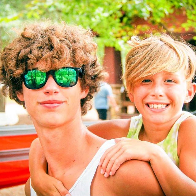 A teenager with curly hair and reflective sunglasses gives a piggyback ride to a smiling young child with side-swept blond hair. Outdoors with trees and a cabin in the background, they're enjoying a sunny day at summer camp. Both are wearing sleeveless shirts.