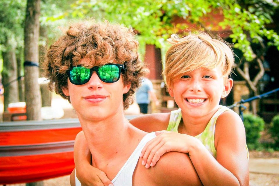 A teenager with curly hair and reflective sunglasses gives a piggyback ride to a smiling young child with side-swept blond hair. Outdoors with trees and a cabin in the background, they're enjoying a sunny day at summer camp. Both are wearing sleeveless shirts.