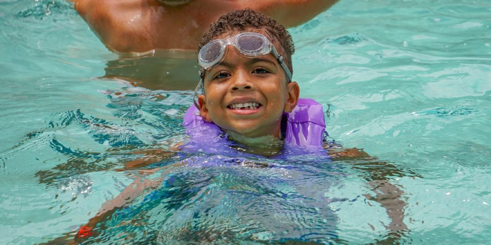 adorable junior camper in life vest smiles in the Camp Canaan pool with goggles on his forehead