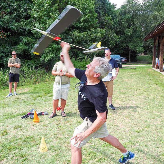 A man is launching a model glider into the air at Camp Canaan during a team building activity, surrounded by trees and a wooden picnic pavilion.