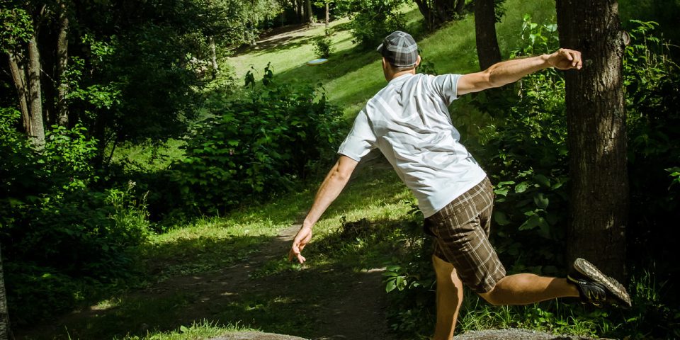 A man wearing a light colored shirt, plaid shorts, and a cap is playing disc golf at Camp Canaan. Mid-throw, with one foot off the ground and his arm extended, the disc soars through the lush, green wooded area—a perfect setting for corporate retreats.