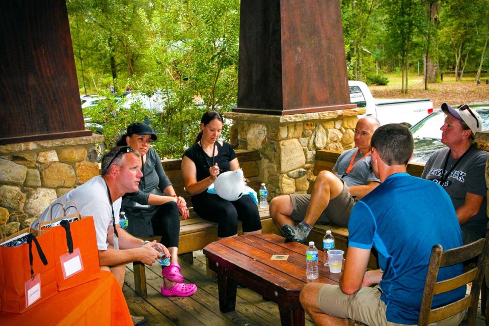 A group of six people sit on the oversized front porch of the multi-purpose building during a team building activity at Camp Canaan.