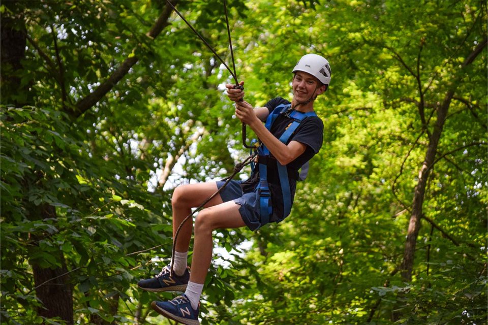 A male camper wearing a helmet and harness is ziplining through a lush forest at Camp Canaan. He is smiling and holding onto the zipline with both hands, enjoying the adventure. The background is filled with green trees and foliage, capturing the essence of nature.