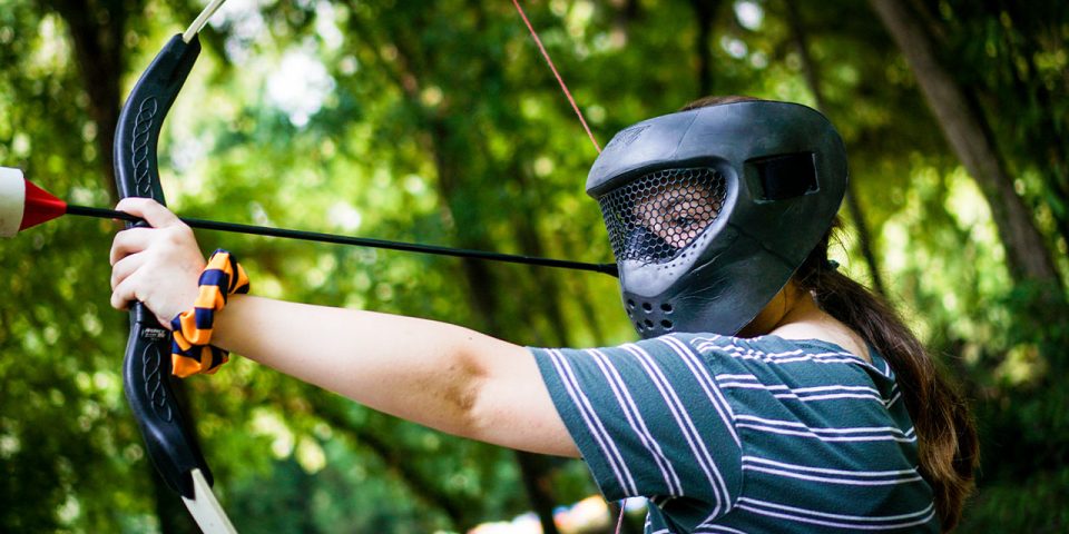 During summer camp, a girl wearing a protective mask aims an archery tag bow and arrow in an outdoor wooded area. Girl is dressed in a green and white striped shirt with a scrunchie on her left wrist.