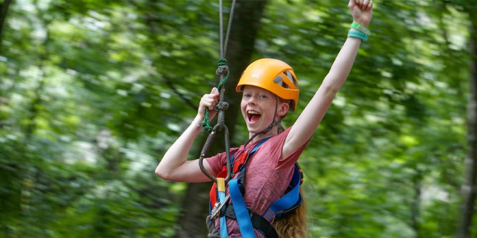 A girl camper wearing an orange helmet and safety harness is ziplining through a wooded area during summer camp.