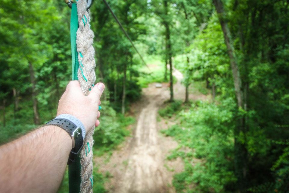 A person wearing a wristwatch and a gray bracelet holds onto a rope on a zipline platform looking down through a lush green forest. The view ahead shows a dirt path winding through tall trees with dense foliage.