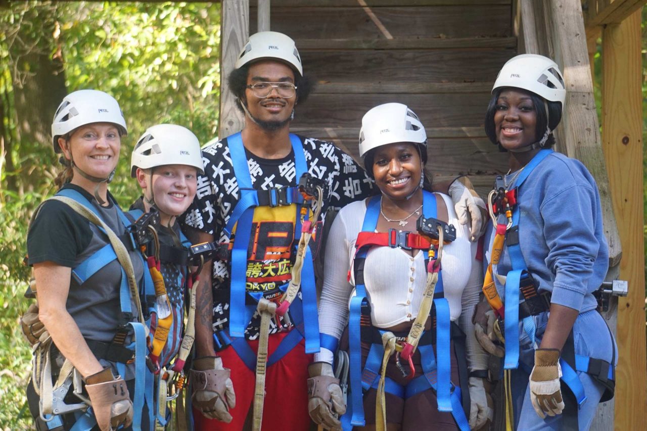 A group of five people wearing white helmets, harnesses, and safety gear stand together, smiling, and posing for a photo. They are about to climb Canaan's zipline canopy tour tower.