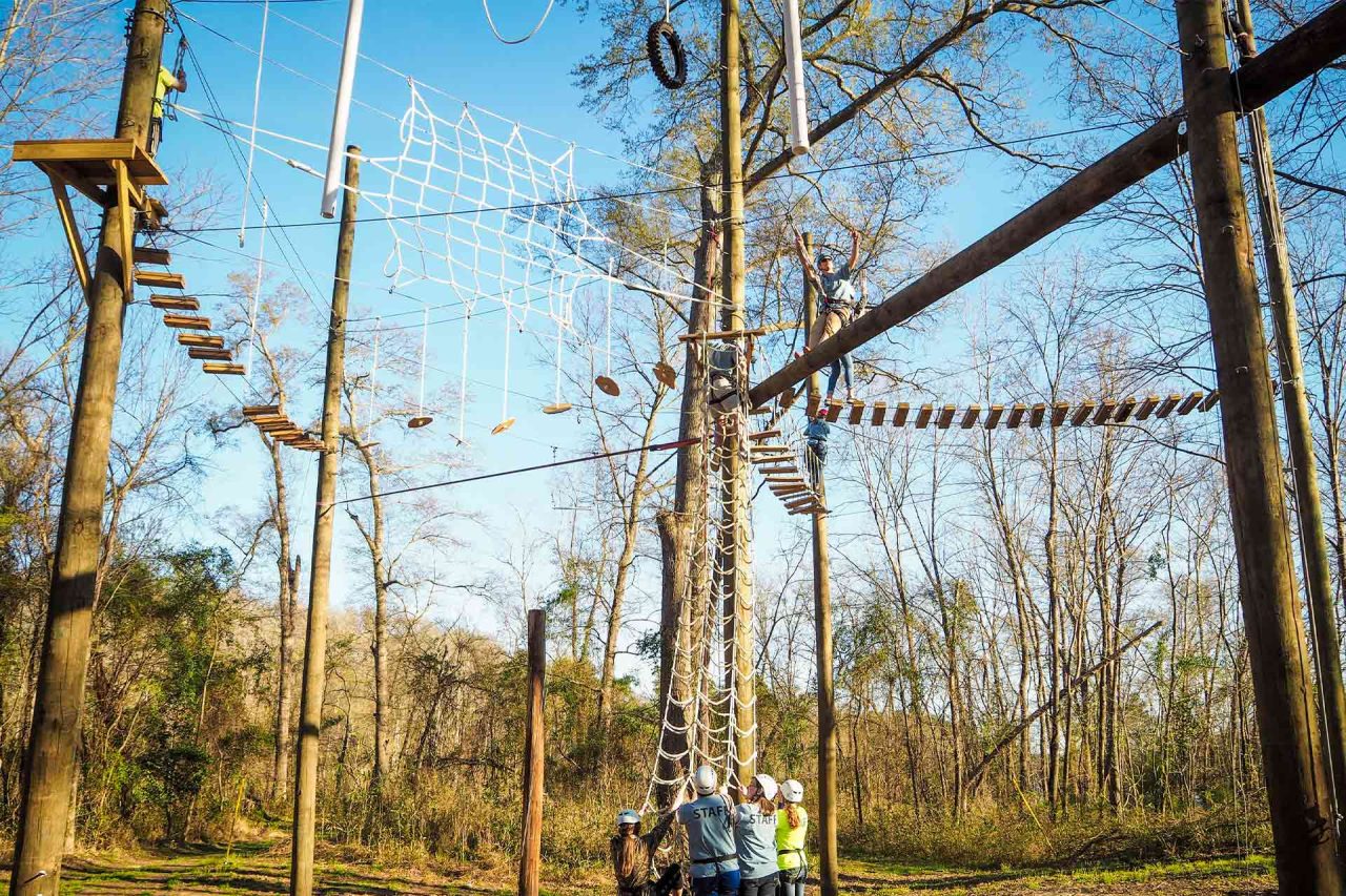 At Camp Canaan, a group of people equipped with helmets gathers at the base of a high ropes course set among trees. The course features rope bridges, swinging elements, and a cargo net, all elevated off the ground. Ideal for summer camp and corporate retreats.