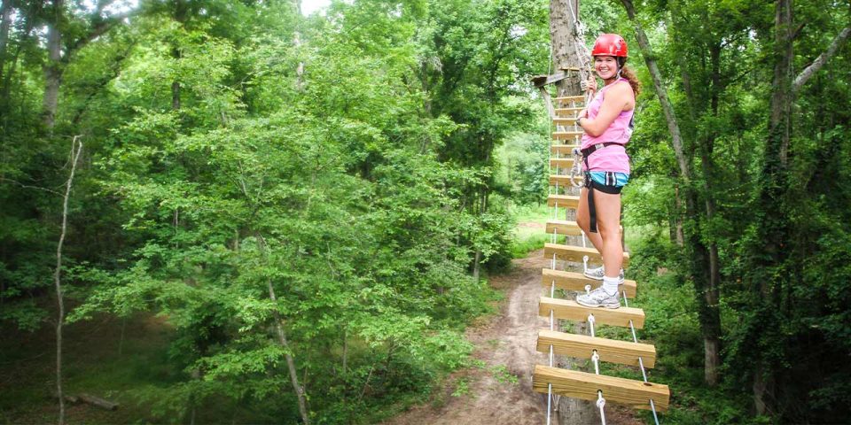 A person wearing a red helmet, pink tank top, and blue shorts is standing on a high ropes course element at Camp Canaan.