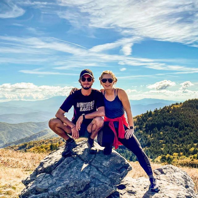 A man and a woman pose on a rocky ledge in a scenic mountainous area under a partly cloudy sky.