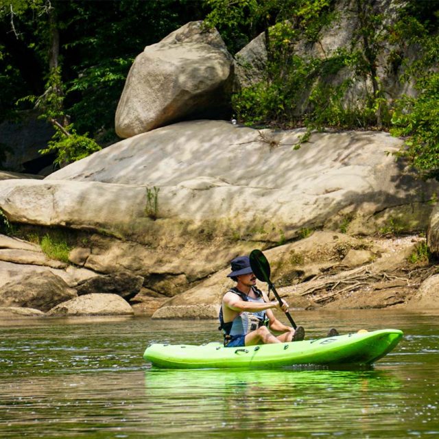 A young man with a mustache wearing a hat paddles a green kayak on the Catawba river surrounded by large rocks and lush greenery, enjoying a peaceful outdoor activity under a sunny sky.