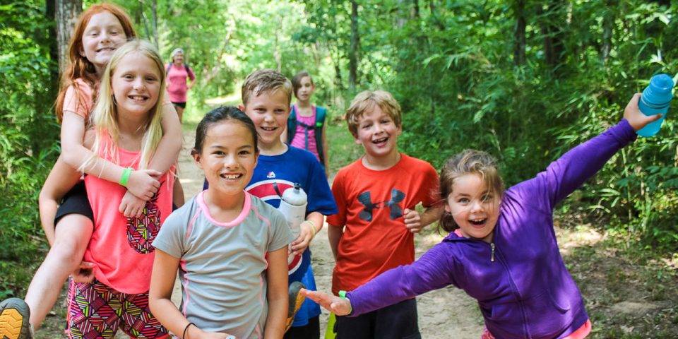 A group of six children happily posing on a sandy path at Camp Canaan. Some are holding water bottles, and one girl is being given a piggyback ride. They are dressed in colorful casual clothes, smiling, and look energetic and joyful. The lush greenery surrounds them during summer camp.