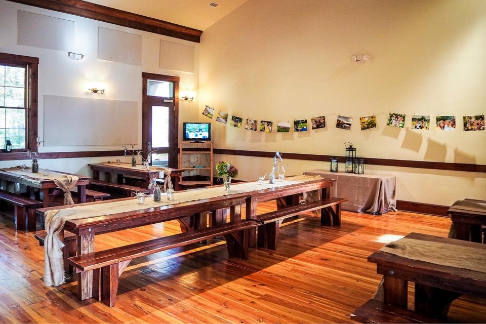 Interior of the multi-purpose building with wooden floors and long wooden benches arranged with simple table settings. A string of photographs hangs on the wall. Soft lighting and decor create a rustic, welcoming atmosphere. There is a table with decor in the corner and a TV on the side wall.