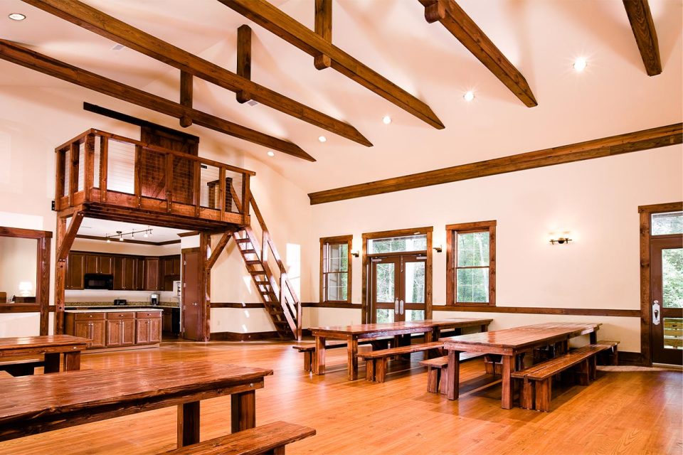 Interior of the multi-purpose building with with wooden beams on the ceiling, wooden floors, and walls with wood trim. It features long wooden tables and benches, a staircase leading to a small loft area, and a kitchenette with cabinets and a sink. Large windows allow natural light to enter.