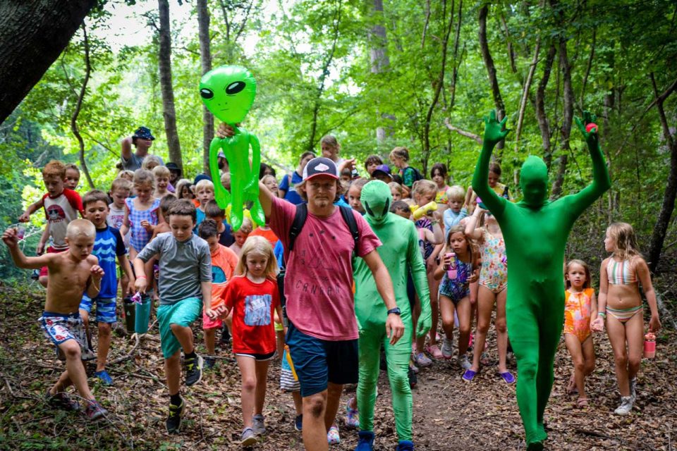 counselor holding inflatable green alien is joined by other counselors dressed as aliens leading junior day campers on a nature walk around Camp Canaan