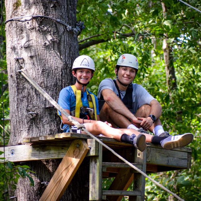 Two people wearing helmets and safety gear sit on a zipline platform attached to a tree, surrounded by lush green foliage.