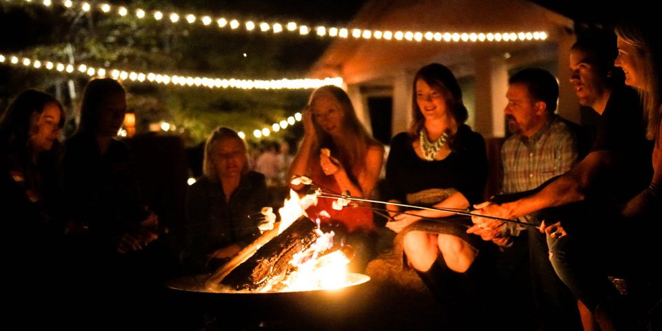 A group of people sit around a campfire at night, roasting marshmallows on sticks. String lights are hung above, adding a warm, festive glow to the scene. Everyone appears engaged and enjoying the moment, with trees and a building faintly visible in the background.