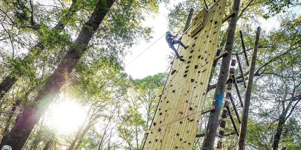 A person climbs the Catawba tower rock climbing wall surrounded by trees with sunlight streaming through the leaves.