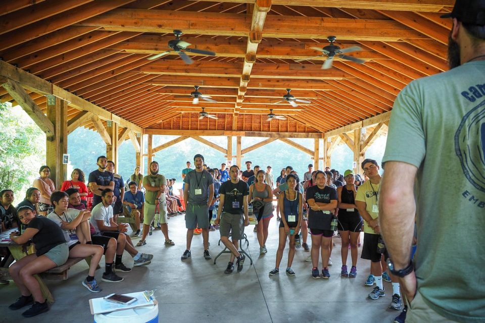 A group of people are gathered under a large wooden picnic pavilion at Camp Canaan. They are paying attention to a person standing at the front, who is speaking to them. Some participants are seated at picnic tables on the left, while others are standing. It appears to be a casual team building activity.