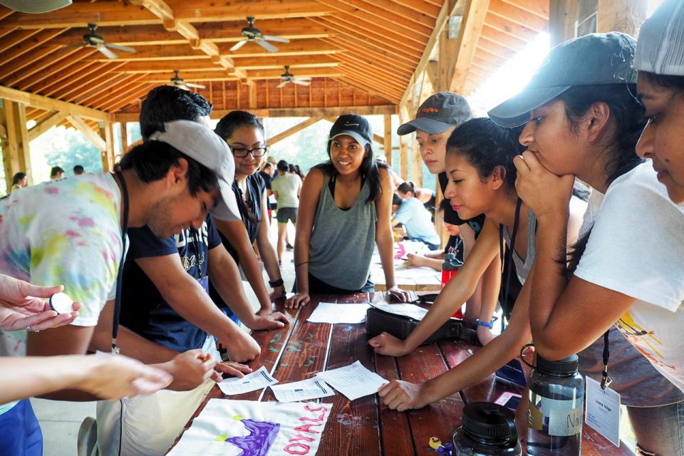 A group of people wearing casual clothes and baseball caps are gathered around a wooden table under the picnic pavilion at Camp Canaan. They are closely looking at papers and items on the table, engaged in a team building activity.