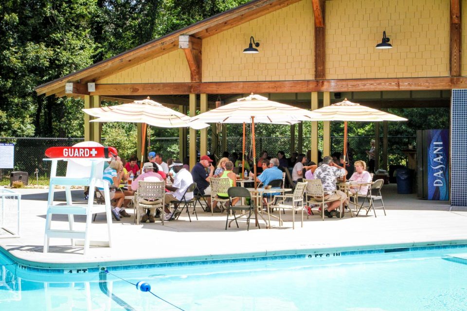 A lively poolside scene features a large group of people seated under umbrellas at tables, engaging in conversation. A life guard station is visible by the pool, which has clear blue water. The backdrop includes a pavilion and trees, with a Dasani vending machine on the side.