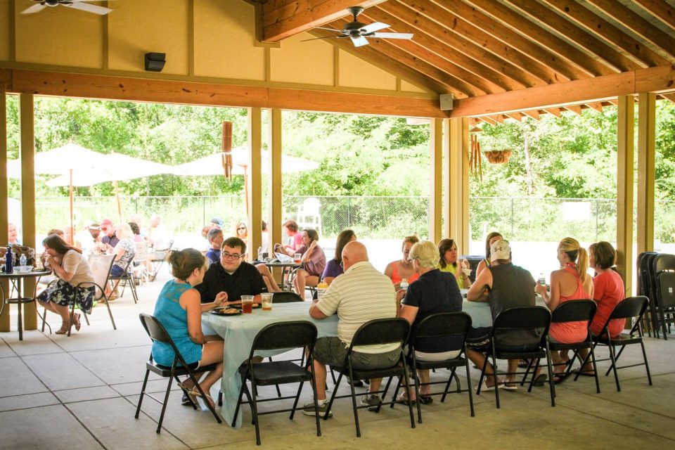 A group of people sit and eat at long tables under an outdoor pavilion situated next to the pool at Camp Canaan.