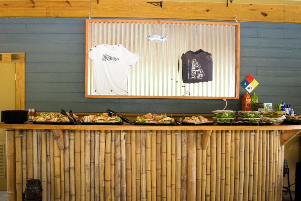 A tiki bar counter is lined with a variety of food dishes, including sandwiches and salads. Above the counter, a corrugated metal backdrop displays two t-shirts: one white with text and one dark with a graphic.